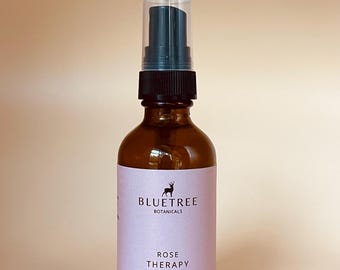 Puede incluir: Un frasco pulverizador de vidrio ámbar de 59 ml con bomba negra y tapa transparente. La etiqueta rosa claro presenta un logotipo de silueta de ciervo, el texto BLUETREE BOTANICALS, ROSE THERAPY y BODY, ROOM AND LINEN MIST.