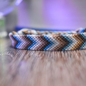 May include: A close-up of a woven friendship bracelet with a chevron pattern in shades of brown, blue, and white.