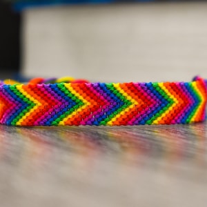 May include: A handmade friendship bracelet with a chevron pattern in vibrant rainbow colors. The bracelet is made of woven threads in red, orange, yellow, green, blue, and purple. The bracelet is resting on a wooden surface.