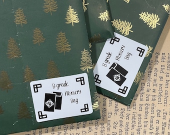 May include: Two white labels with black text and a black illustration of a closed envelope with a red Chinese character on it. The labels are on a green and gold wrapping paper with a pattern of gold pine trees. The text on the labels reads "B-grade Mystery Bag".