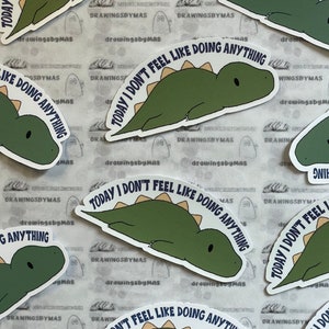 Puede incluir: Una colecci&oacute;n de pegatinas con un dinosaurio verde con una espina dorsal marr&oacute;n claro. El dinosaurio est&aacute; tumbado y el texto "TODAY I DON'T FEEL LIKE DOING ANYTHING" est&aacute; impreso alrededor del dinosaurio. Fondo blanco.
