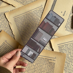 May include: A close-up of a bookmark with a black and white photo of a bookshelf. The bookmark is made of thick cardstock and has a white border. The text "BOOKS BY:" is printed on the bookmark in black.