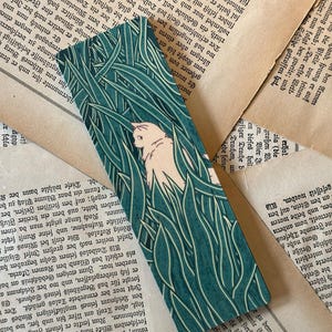 May include: A teal blue bookmark with a white cat and green grass design. The cat is facing left and has a white tail.