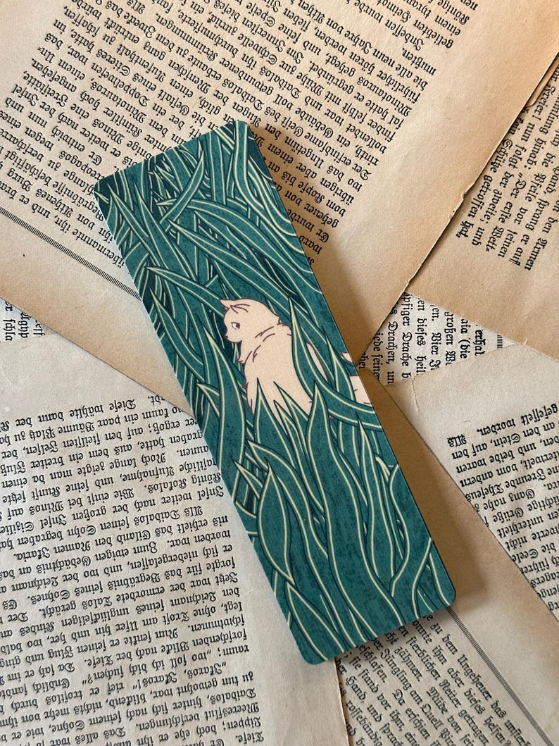 May include: A teal blue bookmark with a white cat and green grass design. The cat is facing left and has a white tail.