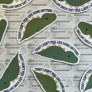 Puede incluir: Una colecci&oacute;n de pegatinas con un dinosaurio de dibujos animados en tonos verdes y beige. Cada pegatina tiene el texto "TODAY I DON'T FEEL LIKE DOING ANYTHING" en una fuente azul oscuro. Las pegatinas est&aacute;n dispersas sobre una superficie estampada.