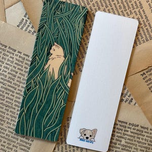 May include: Two bookmarks, one with a green and white pattern of tall grass and a white cat peeking out, and the other with a white background and a cartoon dog peeking out with the text "MAS Library".