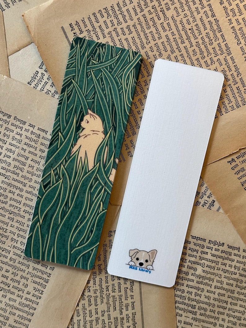 May include: Two bookmarks, one with a green and white pattern of tall grass and a white cat peeking out, and the other with a white background and a cartoon dog peeking out with the text "MAS Library".