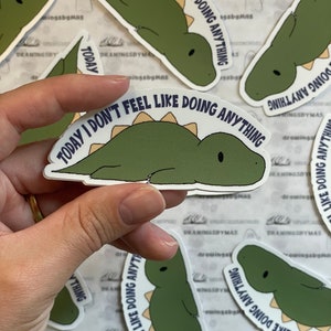 Puede incluir: Una colecci&oacute;n de pegatinas con un dinosaurio verde con el texto "TODAY I DON'T FEEL LIKE DOING ANYTHING". El dinosaurio est&aacute; tumbado con una espina dorsal marr&oacute;n claro. Las pegatinas son blancas y brillantes.