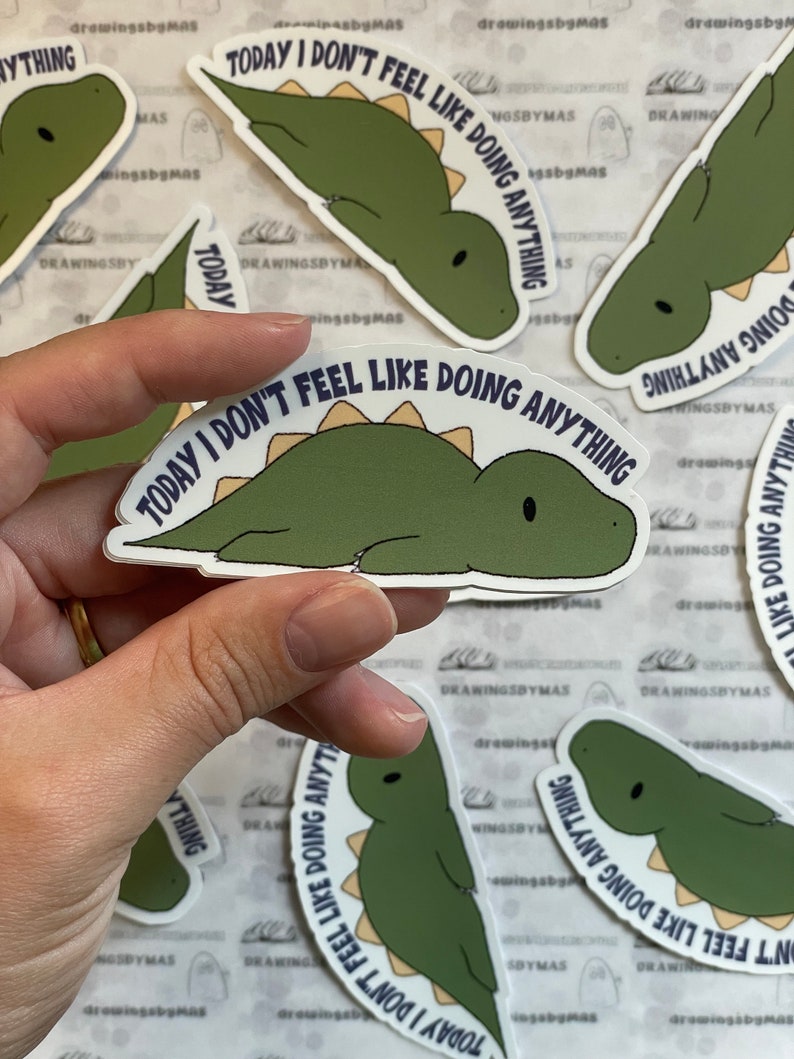 Puede incluir: Una colecci&oacute;n de pegatinas con un dinosaurio verde con el texto "TODAY I DON'T FEEL LIKE DOING ANYTHING". El dinosaurio est&aacute; tumbado con una espina dorsal marr&oacute;n claro. Las pegatinas son blancas y brillantes.