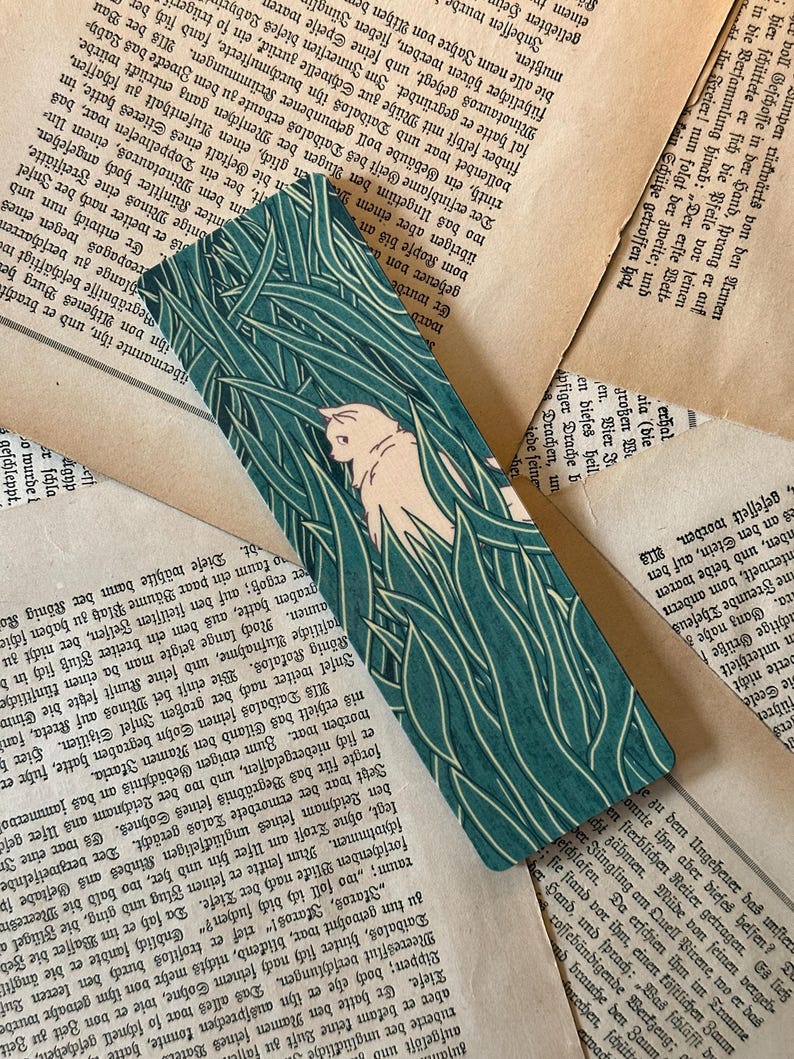 May include: A bookmark featuring a white cat peeking out from behind tall green grass. The cat is facing the right side of the bookmark. The background is a dark green color.
