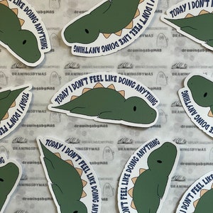 Puede incluir: Un conjunto de pegatinas con un dinosaurio de dibujos animados verde y beige. El texto "TODAY I DON'T FEEL LIKE DOING ANYTHING" en azul marino rodea al dinosaurio. Las pegatinas est&aacute;n sobre un fondo estampado.