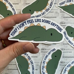 Puede incluir: Una colecci&oacute;n de pegatinas con un dinosaurio de dibujos animados en tonos verdes y beige. El dinosaurio est&aacute; tumbado con el texto "TODAY I DON'T FEEL LIKE DOING ANYTHING" en una fuente curva alrededor del dinosaurio.