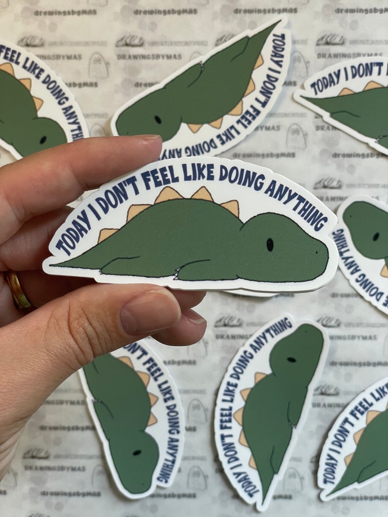 Puede incluir: Una colecci&oacute;n de pegatinas con un dinosaurio de dibujos animados en tonos verdes y beige. El dinosaurio est&aacute; tumbado con el texto "TODAY I DON'T FEEL LIKE DOING ANYTHING" en una fuente curva alrededor del dinosaurio.