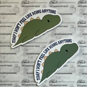 Puede incluir: Dos pegatinas con un dinosaurio verde y espina dorsal beige. El texto "TODAY I DON'T FEEL LIKE DOING ANYTHING" est&aacute; en arco alrededor del dinosaurio. El fondo es una superficie blanca sucia con un patr&oacute;n.