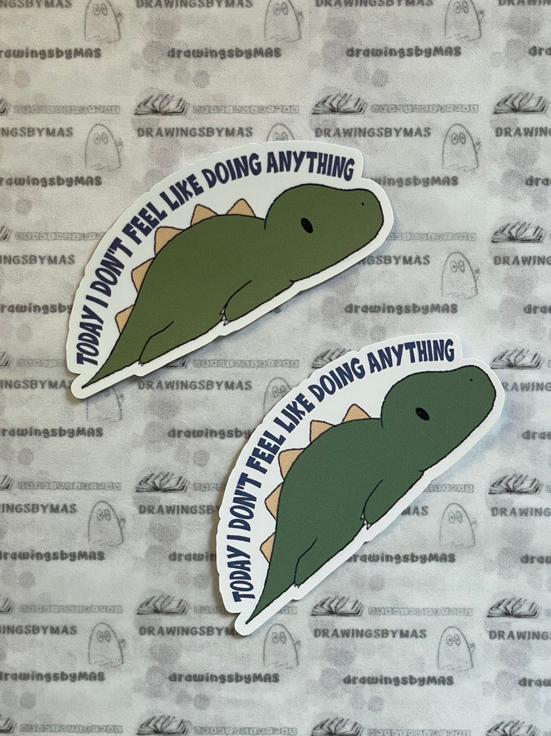 Puede incluir: Dos pegatinas con un dinosaurio verde y espina dorsal beige. El texto "TODAY I DON'T FEEL LIKE DOING ANYTHING" est&aacute; en arco alrededor del dinosaurio. El fondo es una superficie blanca sucia con un patr&oacute;n.