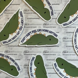 Puede incluir: Un conjunto de pegatinas con un dise&ntilde;o de dinosaurio de dibujos animados en tonos verdes y beige. El texto "TODAY I DON'T FEEL LIKE DOING ANYTHING" est&aacute; escrito en azul oscuro. Las pegatinas est&aacute;n sobre un fondo blanco.
