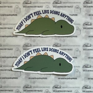 Puede incluir: Dos pegatinas con un dinosaurio verde con espina dorsal beige, tumbado. El texto azul marino "TODAY I DON'T FEEL LIKE DOING ANYTHING" est&aacute; arqueado sobre el dinosaurio. Las pegatinas est&aacute;n sobre un fondo estampado.
