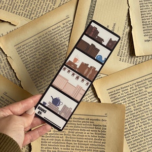 May include: A colorful bookmark with a whimsical design featuring a stack of books, a teapot, a cup of coffee, and a whale. The bookmark has the text "Books By:" at the bottom.