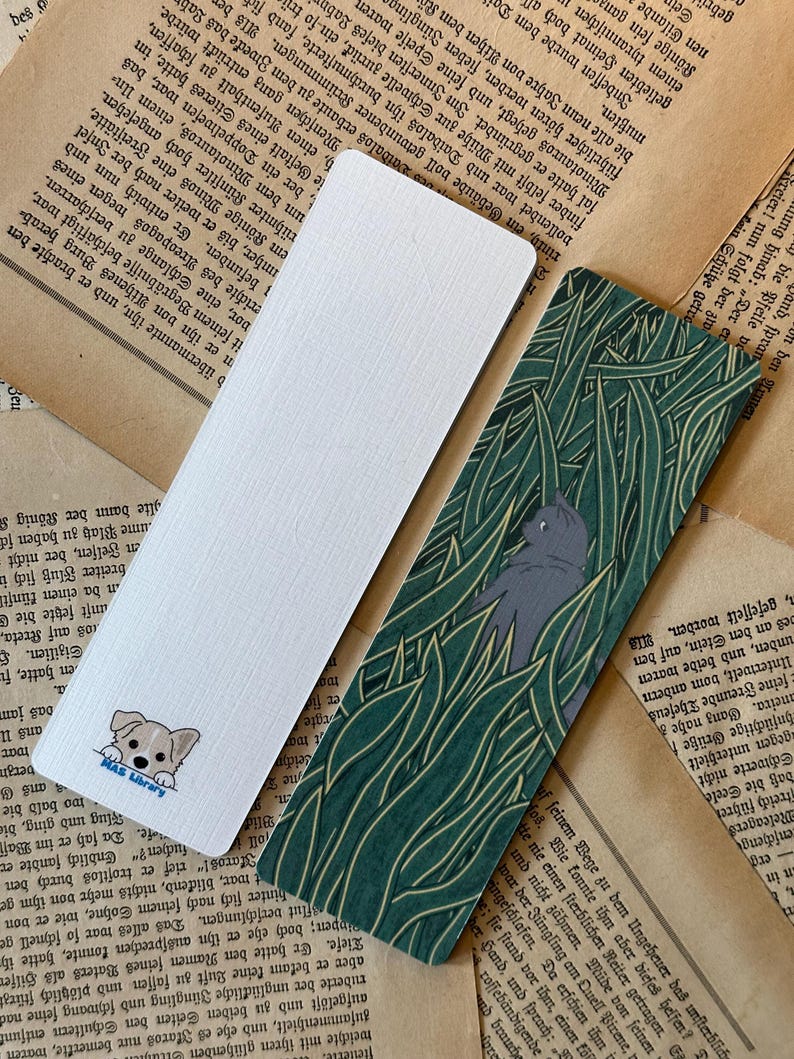 May include: Two bookmarks, one with a white background and a cartoon dog peeking out from the bottom, and the other with a green and yellow leafy pattern and a gray cat peeking out from the top.