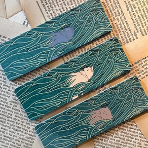 May include: Three colorful bookmarks with a green background and a white outline of tall grass. Each bookmark features a different animal hidden in the grass: a gray cat, a white cat, and a brown cat.