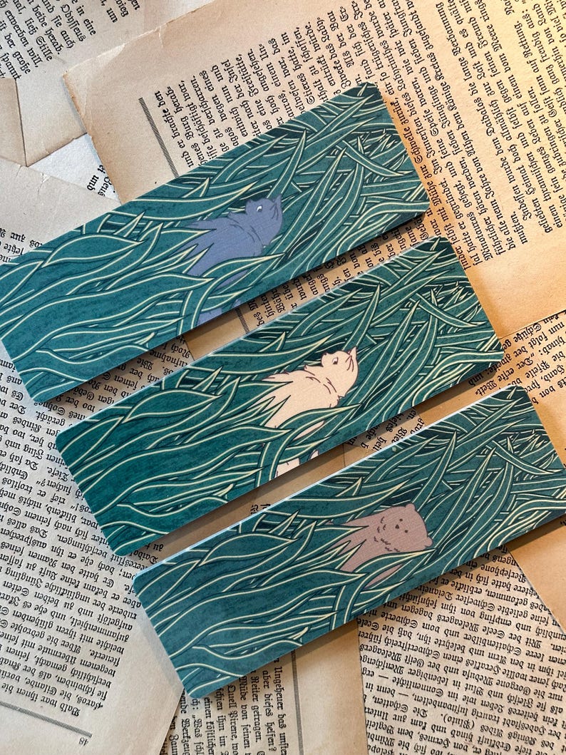 May include: Three colorful bookmarks with a green background and a white outline of tall grass. Each bookmark features a different animal hidden in the grass: a gray cat, a white cat, and a brown cat.