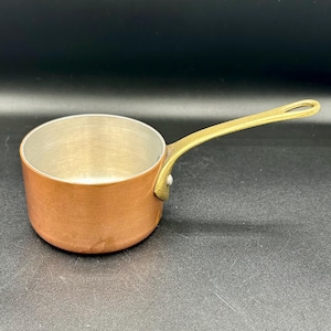 May include: A small saucepan with a polished copper exterior and a silver-toned interior. The cookware features a single long, curved brass handle attached to the side with a visible metal rivet, extending outward from the circular cooking vessel.