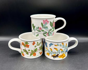 May include: Three white ceramic mugs with floral and butterfly designs. The mugs feature different floral patterns, including pink flowers, orange flowers, and yellow flowers. The mugs have a white background and are decorated with colorful floral and butterfly designs.