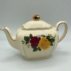May include: A white teapot with a gold lid and handle. The teapot features a floral design with red and yellow roses.