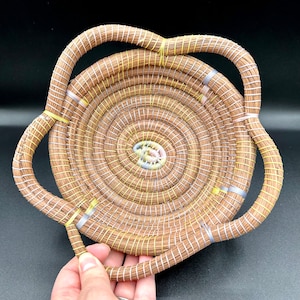 May include: A woven basket with a circular base and two handles. The basket is made of natural materials and has a light brown colour. The handles are woven with yellow and silver accents.