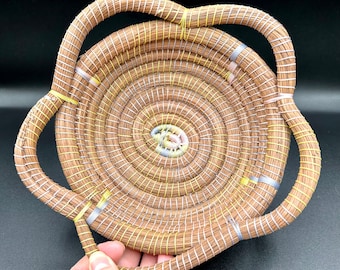 May include: A woven basket with a circular base and two handles. The basket is made of natural materials and has a light brown colour. The handles are woven with yellow and silver accents.