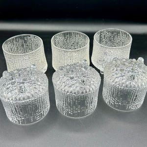 May include: Six clear glass containers. Three are cylindrical with textured vertical lines, and three are round with textured, bubble-like lids. The containers are arranged on a dark surface, showcasing their transparent design.