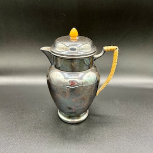 May include: A silver teapot with a yellow knob on the lid and a brown woven handle.