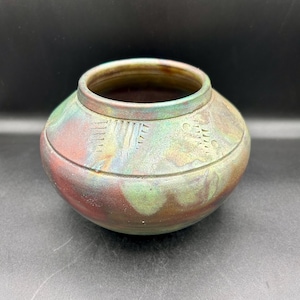 May include: A small, round, ceramic pot with a brown, green and red glaze. The pot has a slightly flared rim and a rounded base. It has a decorative pattern of lines and dots around the middle.