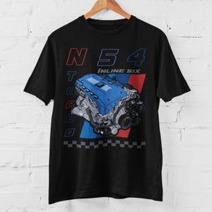 May include: Black t-shirt with a blue and silver engine graphic and the text "N54 Inline Six Turbo". The design includes red and blue stripes and a checkered pattern. The shirt is displayed on a wooden hanger.