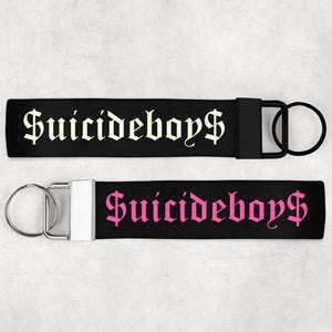 Könnte beinhalten: Zwei schwarze Schlüsselanhänger mit dem Text "Suicideboys$" in gotischer Schrift. Einer hat neongelben Text, der andere rosa. Beide haben einen Metall-Schlüsselring. Geeignet für Fans.