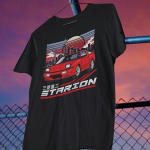 May include: Black t-shirt with a red car graphic and the text "STARION" and Japanese characters. The car is set against a city skyline with a sunset in the background.