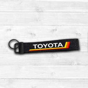 May include: Black keychain with the word "TOYOTA" in white, and a yellow, orange, and red stripe design. The keychain has a metal ring and clasp. The background is a white wooden surface.