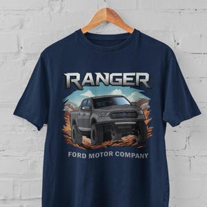Può includere: Una t-shirt blu navy con una grafica di un pick-up Ford Ranger grigio che guida attraverso un paesaggio desertico. Il testo "RANGER" è sopra il camion e "FORD MOTOR COMPANY" è sotto il camion.