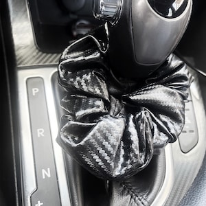以下が含まれることがあります： カーボンファイバー模様の黒いシュシュが車のギアシフトに巻き付けられています。シュシュは光沢のある質感のある素材でできており、束ねられてボリューム感を出しています。ギアシフトは黒とシルバーです。