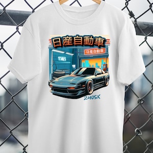 Puede incluir: Camiseta blanca con un gráfico de un Nissan 240SX negro frente a un taller de automóviles japonés con el texto "日産自動車" (Nissan Automobile) y "240SX".