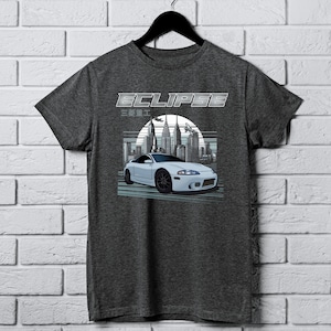 Puede incluir: Una camiseta gris oscuro con un gráfico de coche blanco y el texto "ECLIPSE" en fuente plateada. El coche es un Mitsubishi Eclipse blanco con un horizonte urbano en el fondo. El texto "三 菱 ｴｸﾘﾌﾟｽ" también está impreso en la camiseta.