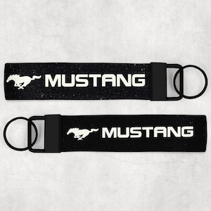 May include: Two black keyrings with the word "MUSTANG" in white block letters. Each keyring has a white running horse logo and a black metal ring. The keyrings are displayed on a light grey background.