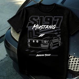 Może przedstawiać: Czarna koszulka z grafiką czarnego samochodu Mustang, napisem "S197 Mustang" i "American Muscle". Projekt zawiera stylizowaną amerykańską flagę.