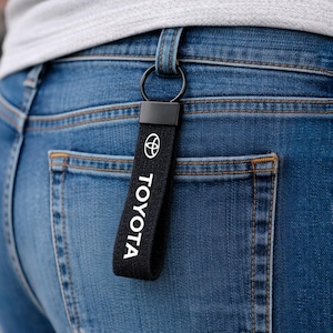 May include: Black Toyota keyring with white logo and text, attached to a black metal ring. The keyring is hanging from a belt loop on blue denim jeans.