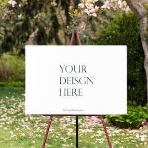 May include: A white rectangular sign on a brown easel, with the text "YOUR DESIGN HERE" in navy blue. The sign is set outdoors in a grassy area with trees and fallen flower petals in the background.