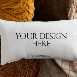 Puede incluir: Almohada blanca con el texto "YOUR DESIGN HERE" y el texto "OnTrendMockupCo." sobre una superficie de madera marrón con una cesta tejida, una almohada amarilla y una manta de cuadros blancos y naranjas.