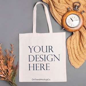 Puede incluir: Una bolsa de tela blanca con el texto "YOUR DESIGN HERE" y el sitio web "OnTrendMockupCo." impreso en ella. La bolsa está sobre una superficie gris con una bufanda de punto marrón y un reloj vintage.
