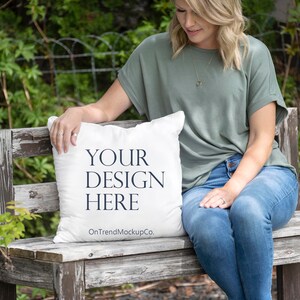 May include: A white pillow with the text "YOUR DESIGN HERE" and the words "OnTrendMockupCo." printed on it. The pillow is sitting on a wooden bench with a woman sitting on the bench.