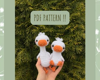May include: Two white crocheted duck plushies with orange beaks and black eyes. The ducks are being held in a person's hands. The text "PDF PATTERN!!" is in a white rectangle with a brown outline.
