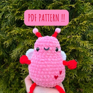 Könnte beinhalten: Ein rosa gehäkeltes Amigurumi-Spielzeug mit roten Armen und Beinen, weißen Flügeln und zwei roten Herzen auf dem Rücken. Das Spielzeug hat ein lächelndes Gesicht mit schwarzen Augen und einem schwarzen Mund. Der Text "PDF PATTERN!!" ist in Rot auf rosa Hintergrund geschrieben.